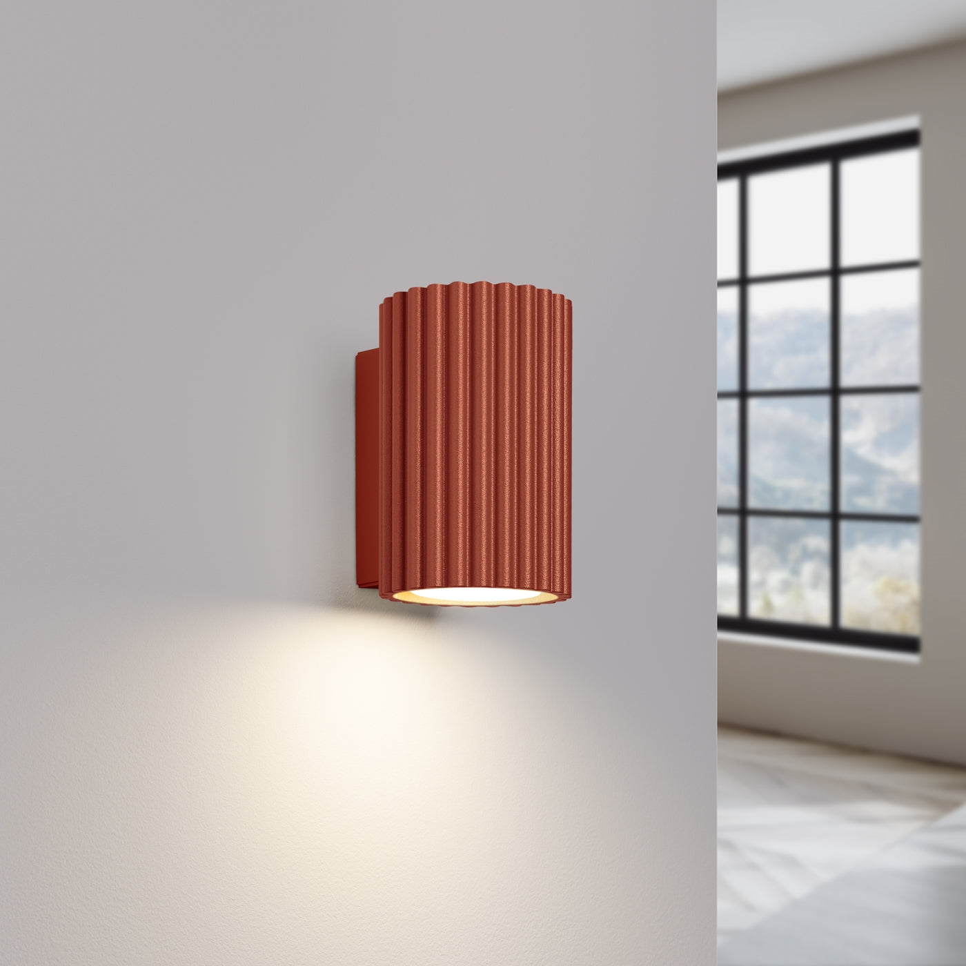 A modern wall sconce with a ribbed terracotta shade, emitting a warm light, mounted on a light gray wall. A large window in the background allows natural light into the space.