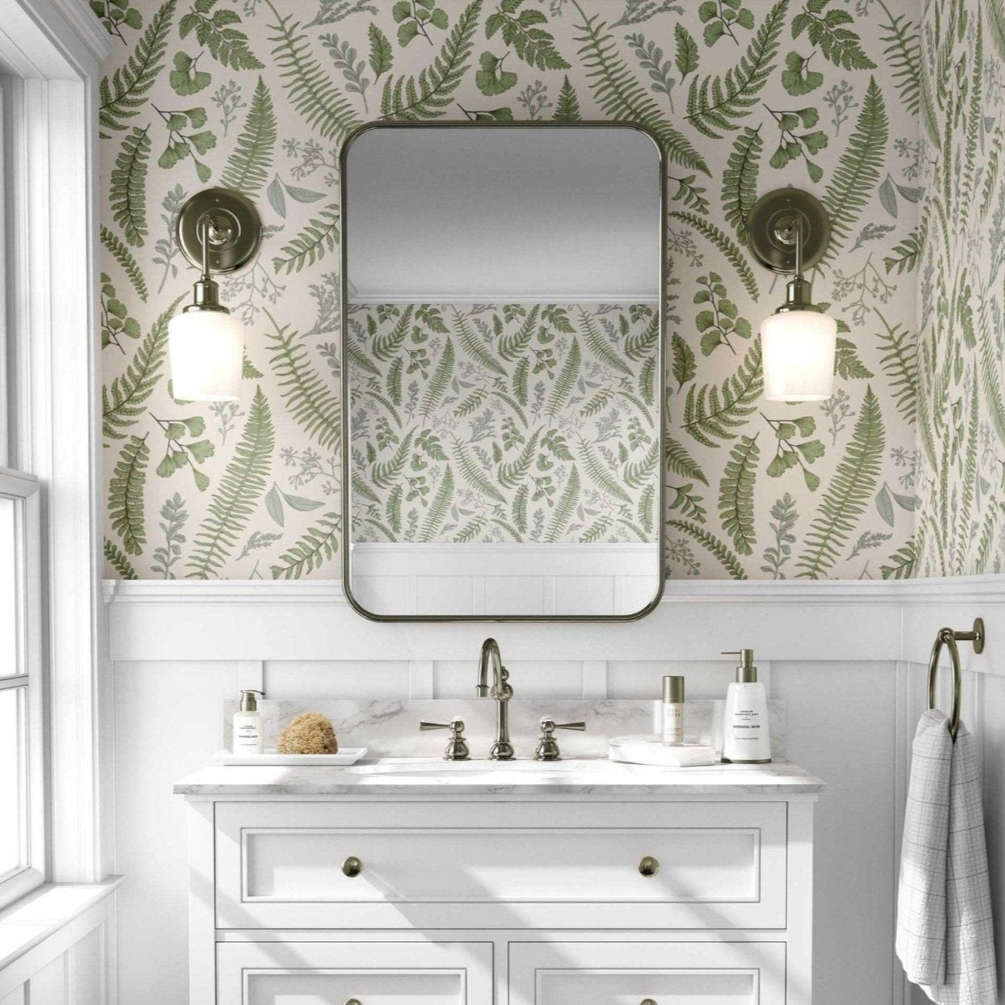 Bathroom with green fern leaves with a beige background wallpaper and a white vanity with gold accents. A large rectangular mirror with rounded edges hangs above the vanity, flanked by two brass sconces with white globe lights.