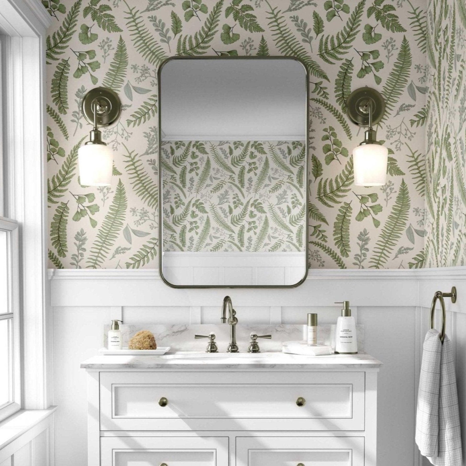 Bathroom with green fern leaves with a beige background wallpaper and a white vanity with gold accents. A large rectangular mirror with rounded edges hangs above the vanity, flanked by two brass sconces with white globe lights.