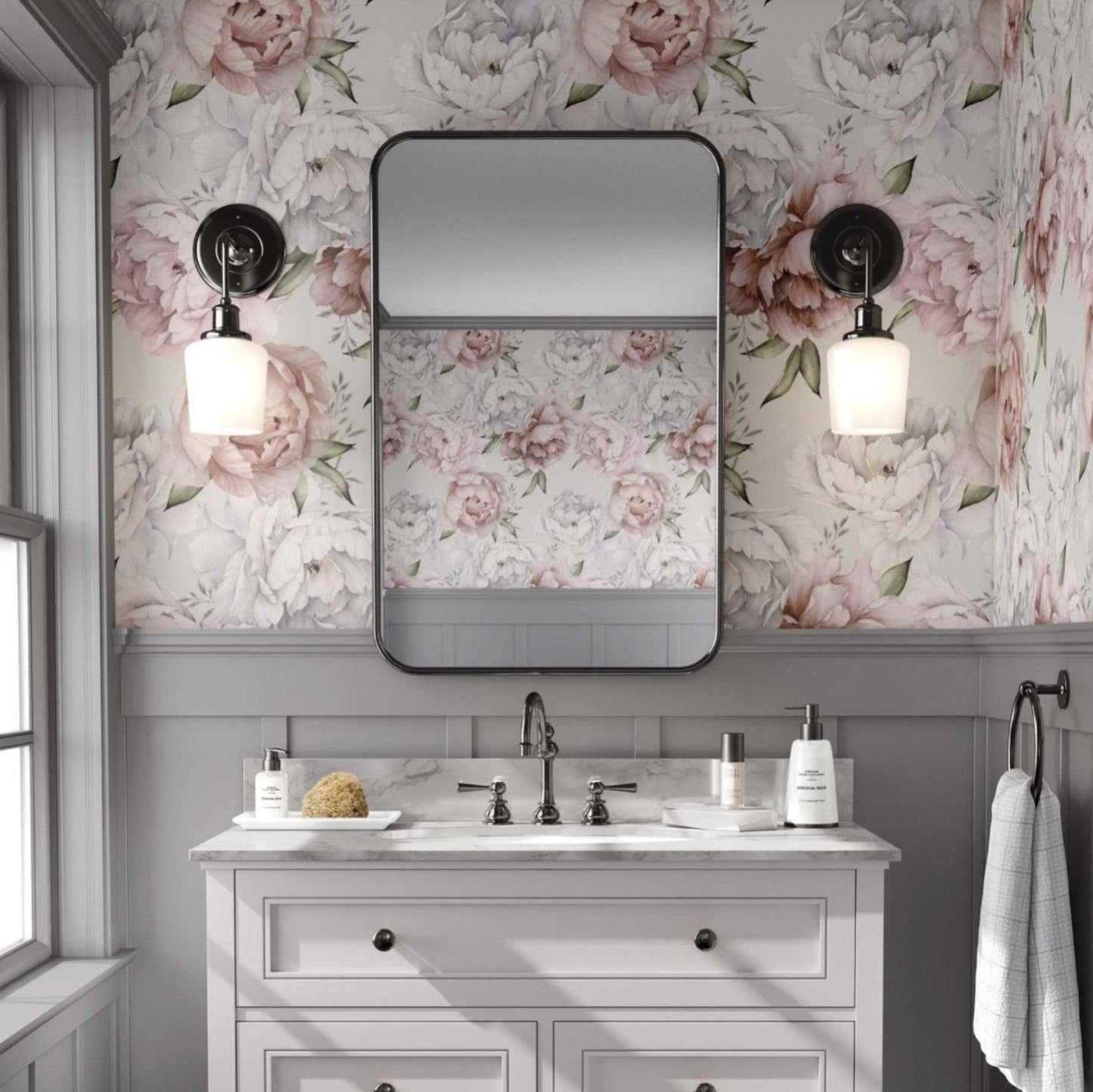 Bathroom with a light pink and white peony floral wallpaper, featuring large flowers against a beige background. A white vanity with black fixtures stands below a rectangular mirror with rounded edges, flanked by brass sconces with globe lights. The space has an elegant, vintage feel, with grey paneling beneath the wallpaper and a window to the left allowing natural light.