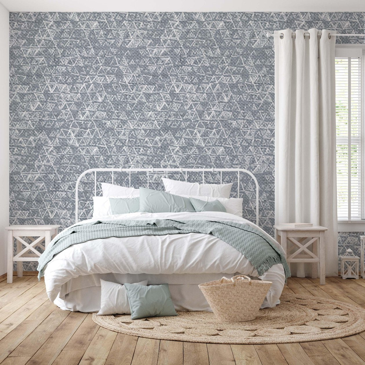 edroom featuring a bold grey wallpaper with an intricate geometric triangular pattern. The wallpaper creates a stylish backdrop behind a white metal bed frame with soft pastel bedding. The room is decorated with light wooden furniture, white curtains, and a woven rug, creating a cozy and modern atmosphere.