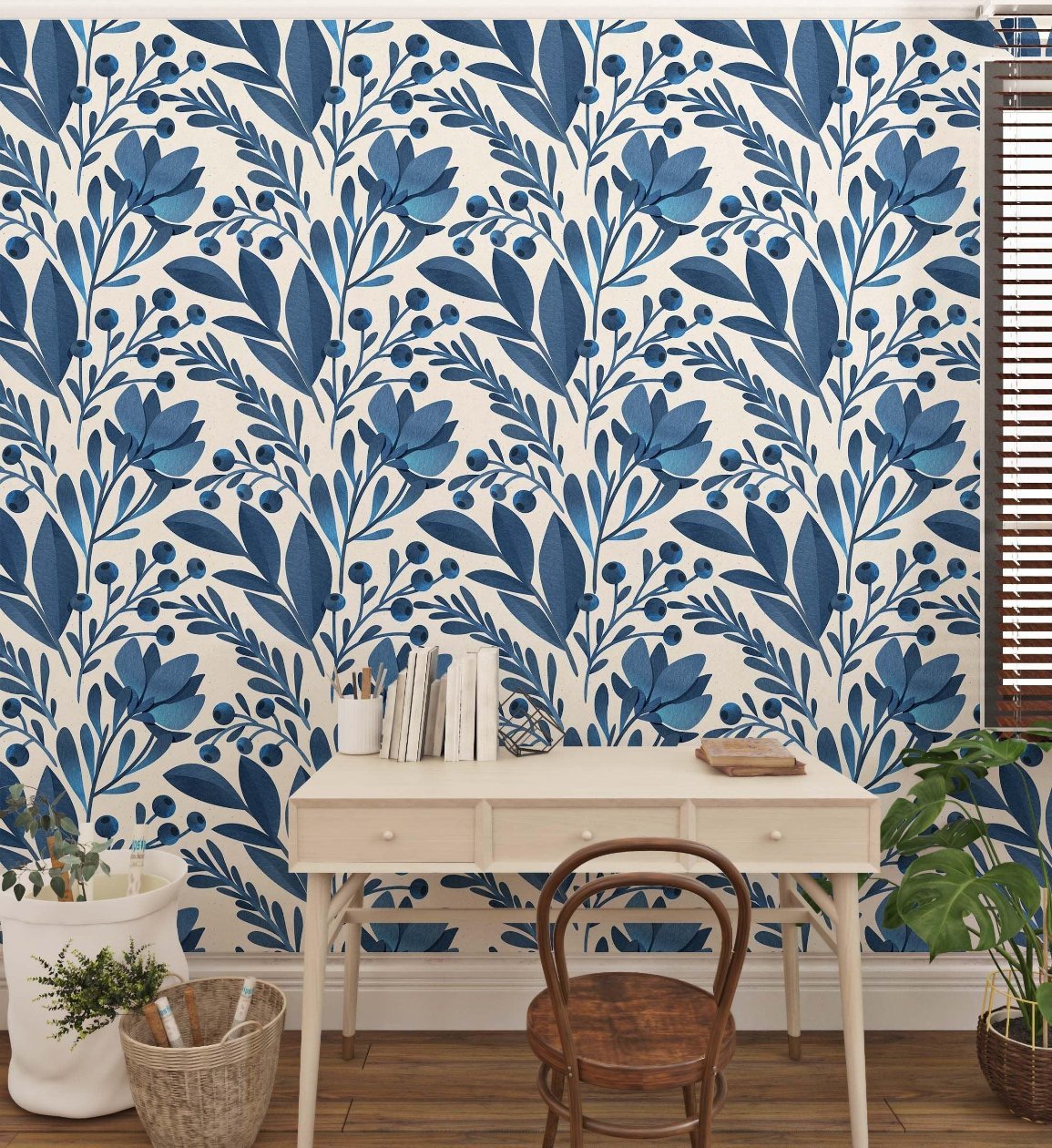 A charming workspace featuring a wooden desk with a light finish and a simple chair. The background showcases vibrant blue floral-patterned wallpaper, adding a fresh, nature-inspired feel to the room. A few houseplants and a basket with rolled-up papers enhance the cozy ambiance.