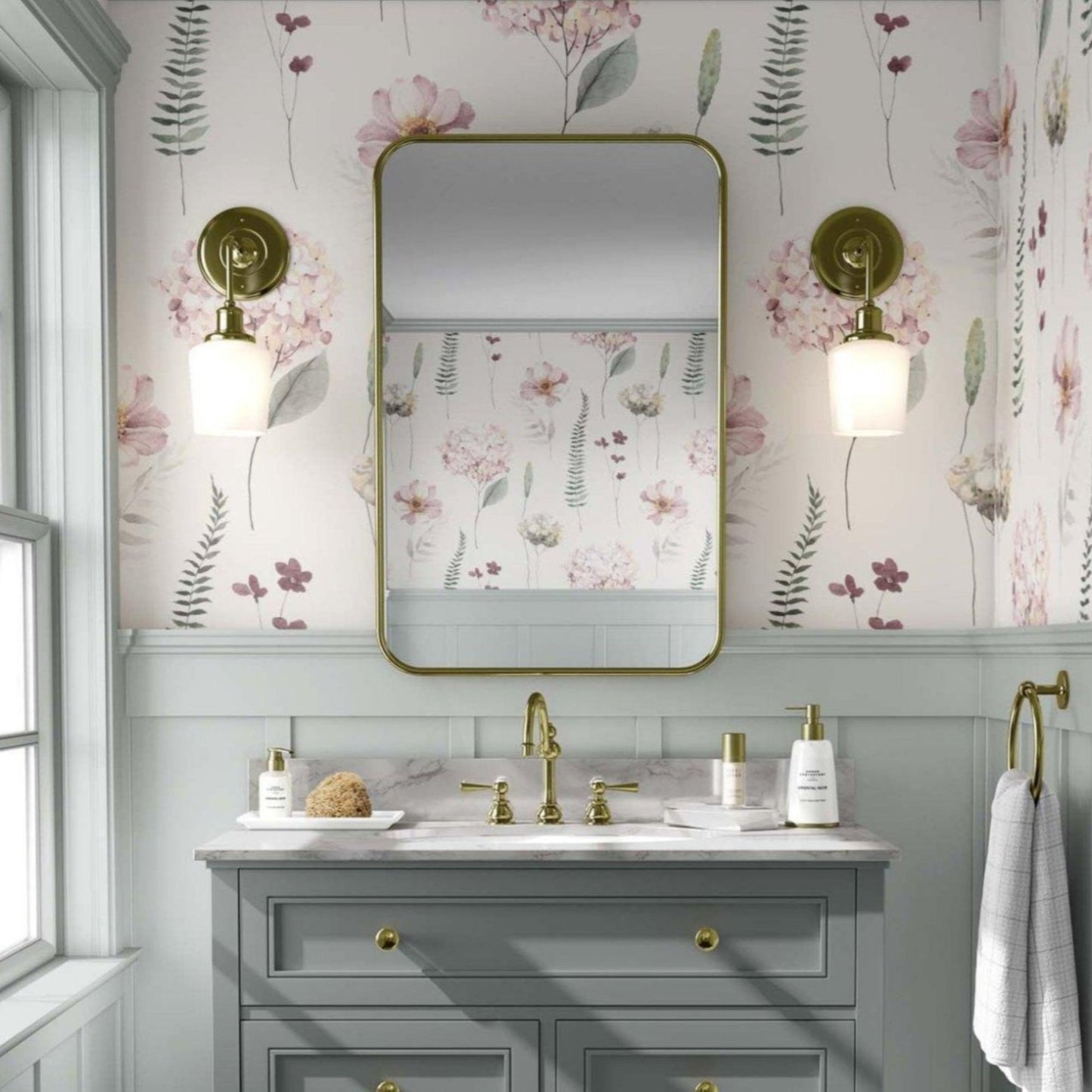 Bathroom with light pink hydrangea flowers and greens floral wallpaper. A white vanity with gold fixtures stands below a rectangular mirror with rounded edges, flanked by brass sconces with globe lights. The space has an elegant, vintage feel, with light grey paneling beneath the wallpaper and a window to the left allowing natural light.