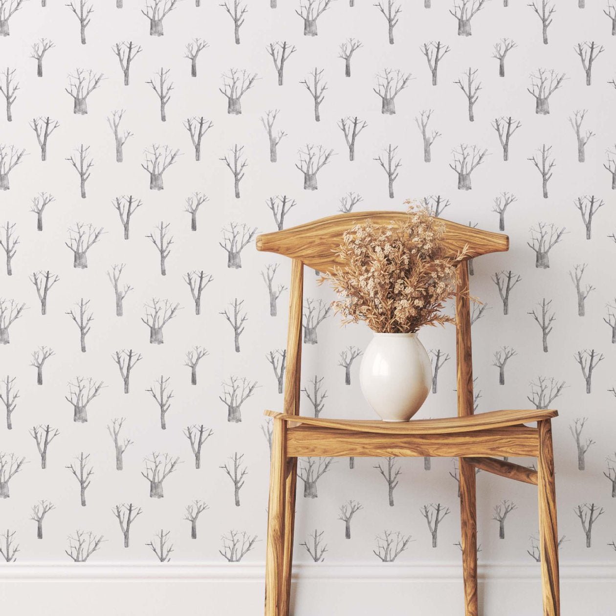 A stylish wooden chair with a natural finish is placed against a wall adorned with subtle, minimalist tree-patterned wallpaper in gray tones. A white vase filled with dried branches sits on the chair, adding a rustic touch to the simple yet elegant decor.