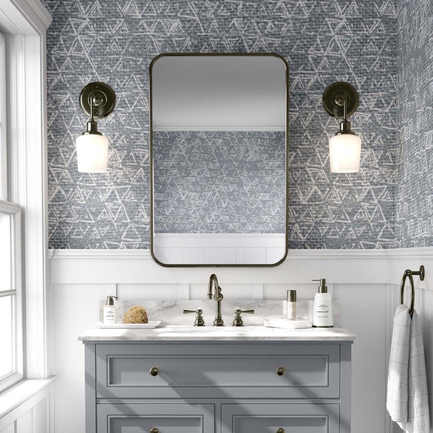 Bathroom with a a bold grey wallpaper with an intricate geometric triangular pattern. A light grey vanity with gold fixtures stands below a rectangular mirror with rounded edges, flanked by brass sconces with globe lights. The space has an elegant, vintage feel, with white paneling beneath the wallpaper and a window to the left allowing natural light.