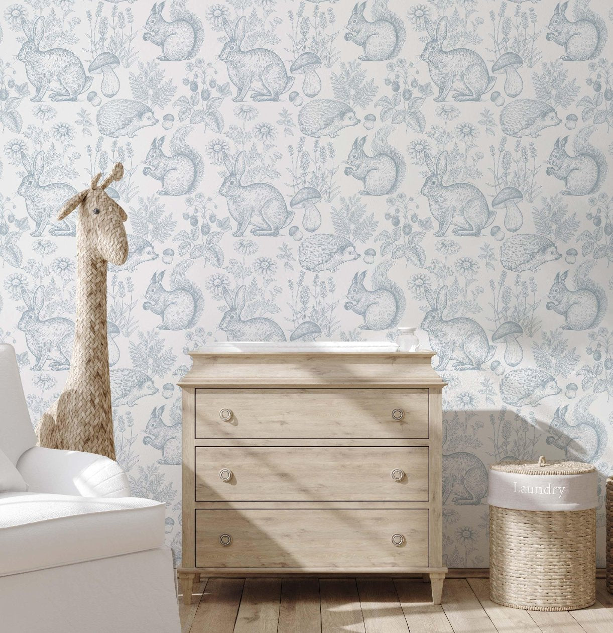 Charming nursery with a wooden chest of drawers, a white arm chair, and a soft woodland-themed wallpaper featuring hand-drawn animals such as squirrels, rabbits, and hedgehogs. The room includes a light wood dresser and a small shelf displaying decorative pieces like animal figurines, adding a cozy, storybook touch."