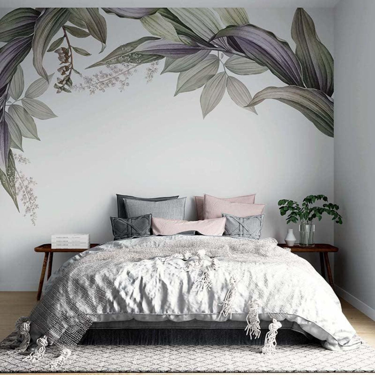 Stylish bedroom with a bed draped in a grey, fringed blanket and layered pillows in soft pink and charcoal grey. The room is accented by a leafy wall mural of oversized green and purple foliage, while simple wooden side tables hold books, a plant, and a vase.