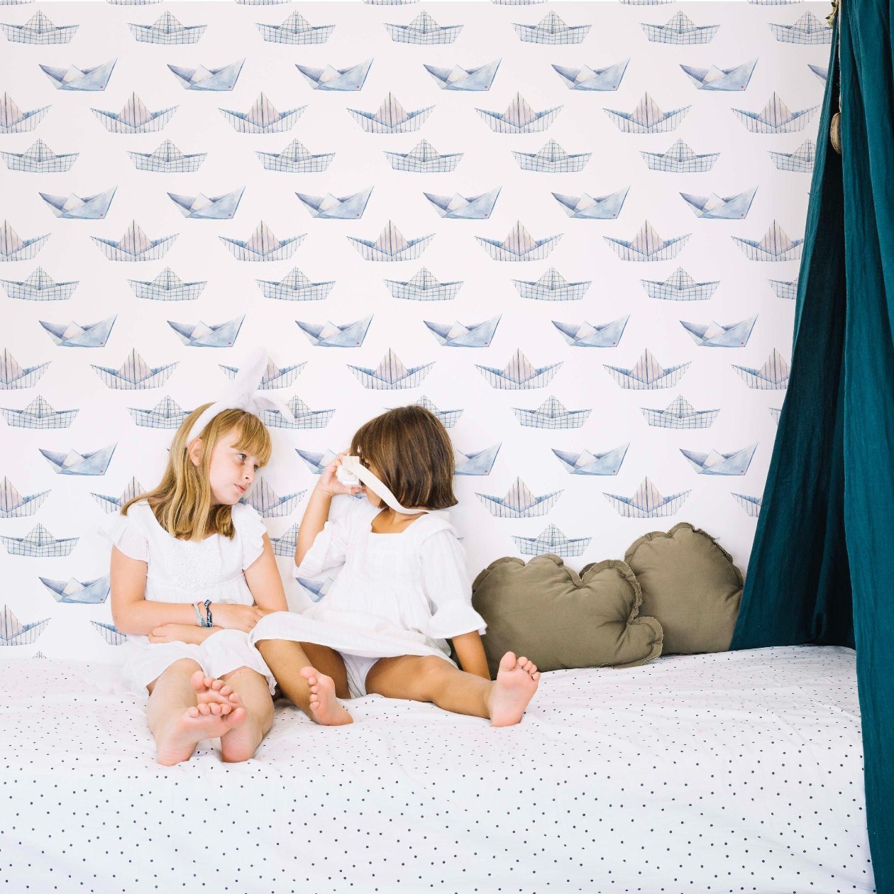 Two girls on a bed, smiling and chatting, capturing a joyful moment of friendship and connection in kids bedroom featuring a paper boat wallpaper in blue.