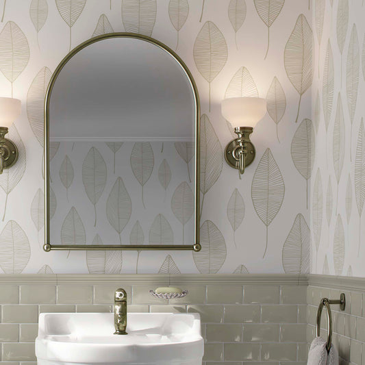 Bathroom interior featuring a minimalist beige leaf wallpaper with a delicate linear pattern, paired with light olive green subway tiles, a large arched brass mirror, and elegant wall sconces.