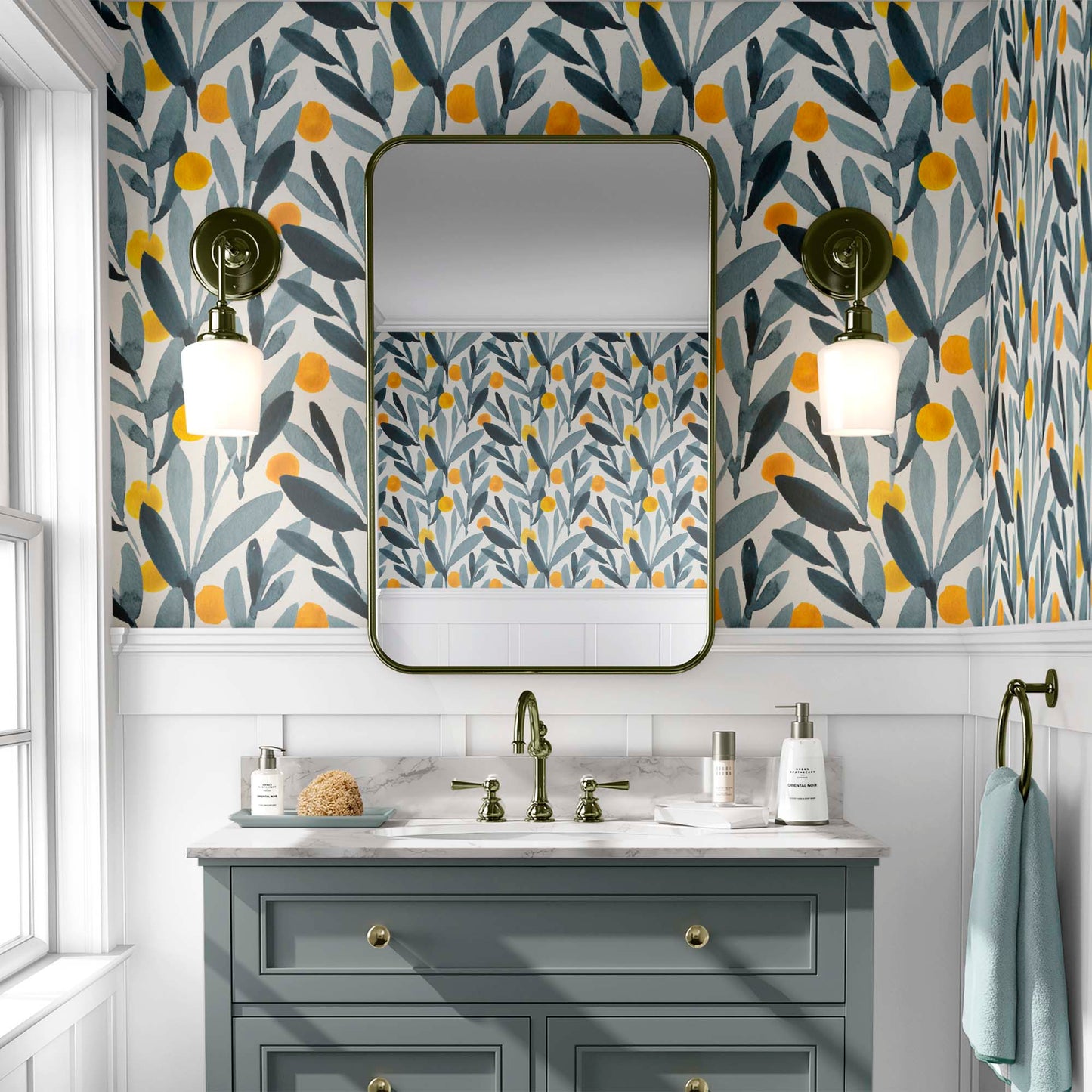 A bathroom featuring wallpaper with a watercolor design of yellow citrus-like shapes and blue-green leaves. The room includes a dark-framed rectangular mirror, wall sconces with white shades, and a sage-green vanity with a marble countertop and brass hardware.
