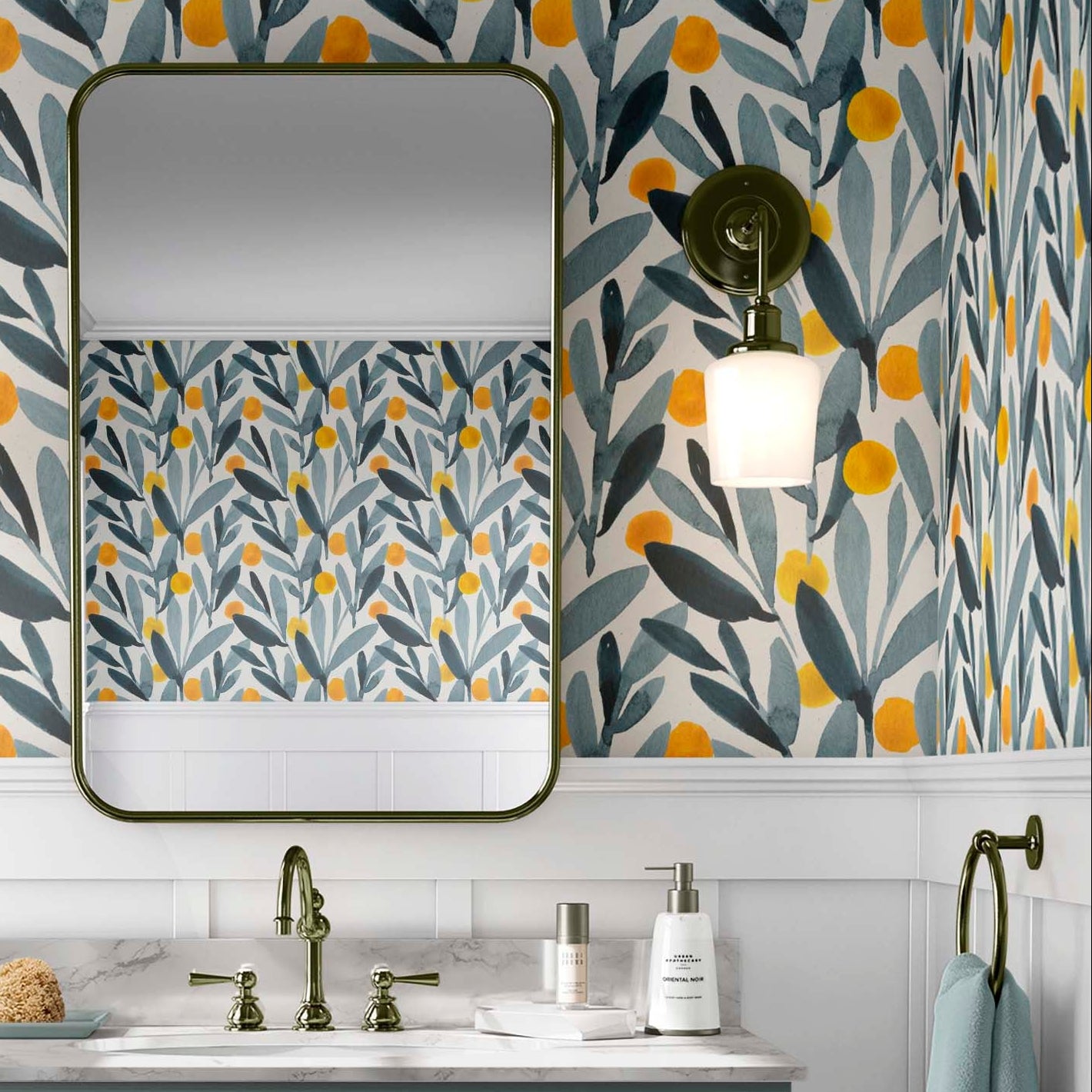 cat-610804236616 A bathroom featuring wallpaper with a watercolor design of yellow citrus-like shapes and blue-green leaves. The room includes a dark-framed rectangular mirror, wall sconces with white shades, and a sage-green vanity with a marble countertop and brass hardware.