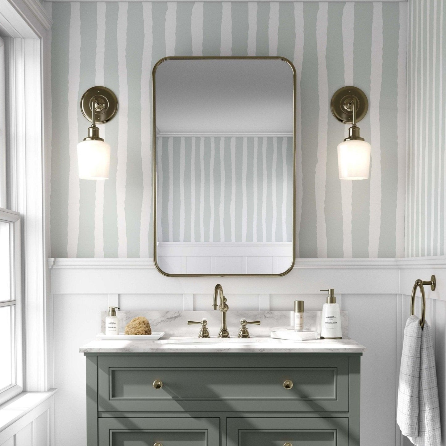 Bathroom with a mint green and white striped wallpaper, featuring irregular stripes. A sage green vanity with gold fixtures stands below a rectangular mirror with rounded edges, flanked by brass sconces with globe lights. The space has an elegant, vintage feel, with white paneling beneath the wallpaper and a window to the left allowing natural light.