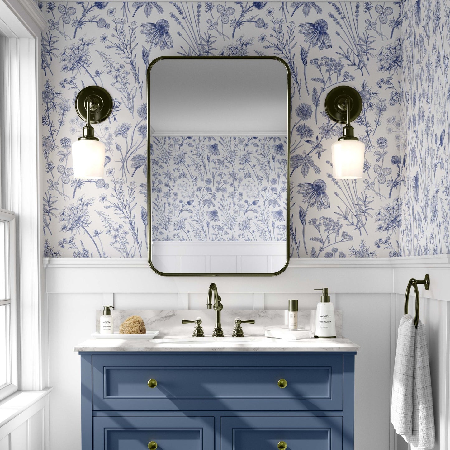 cat-610804236616 A brightly lit bathroom featuring blue and white botanical wallpaper with detailed sketches of wildflowers and meadow plants. The pattern is paired with classic white wainscoting and a slate blue vanity with gold hardware. A rectangular mirror with rounded corners is centered between two antique-style brass wall sconces, reflecting the floral pattern on the opposite wall.