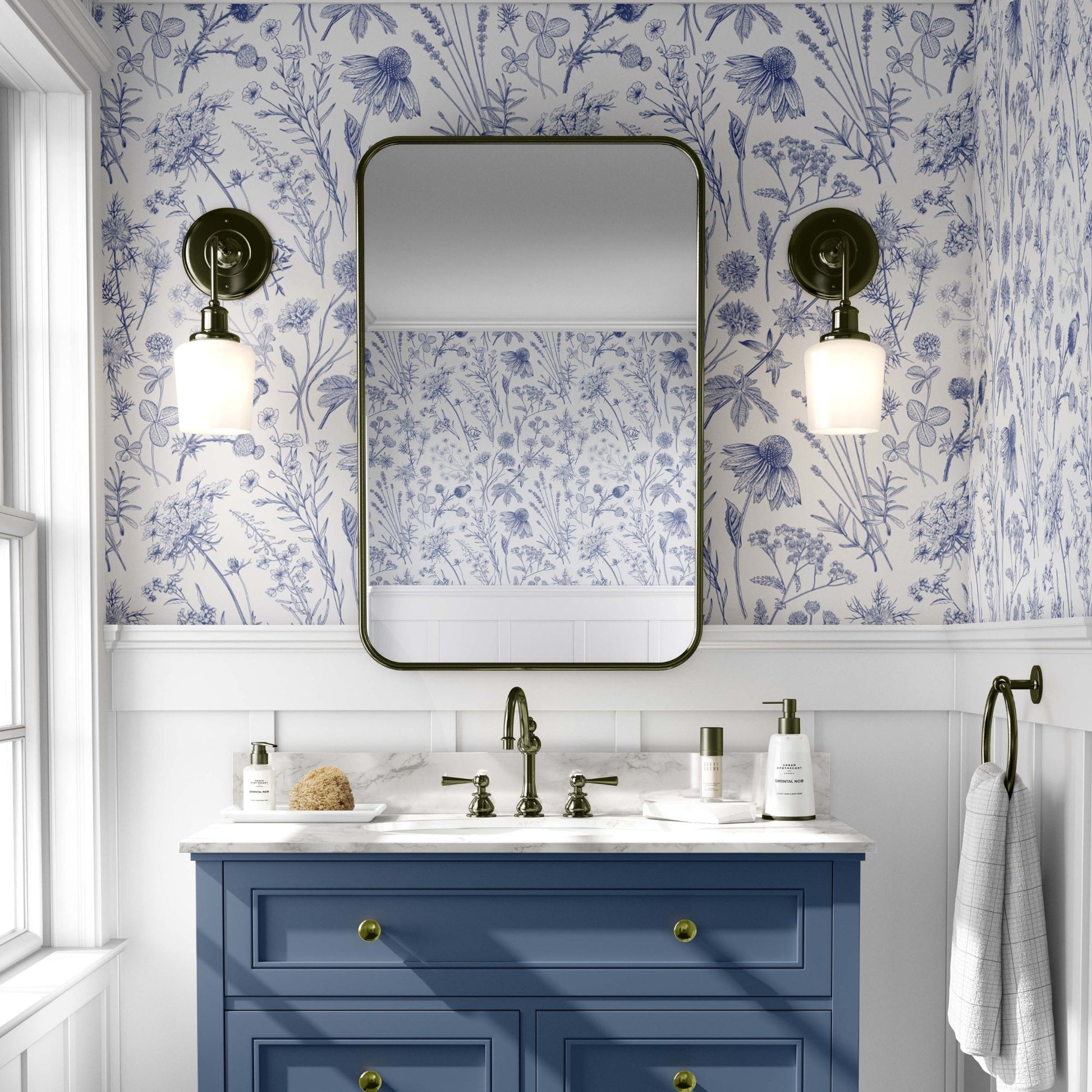 cat-610804236616 A brightly lit bathroom featuring blue and white botanical wallpaper with detailed sketches of wildflowers and meadow plants. The pattern is paired with classic white wainscoting and a slate blue vanity with gold hardware. A rectangular mirror with rounded corners is centered between two antique-style brass wall sconces, reflecting the floral pattern on the opposite wall.
