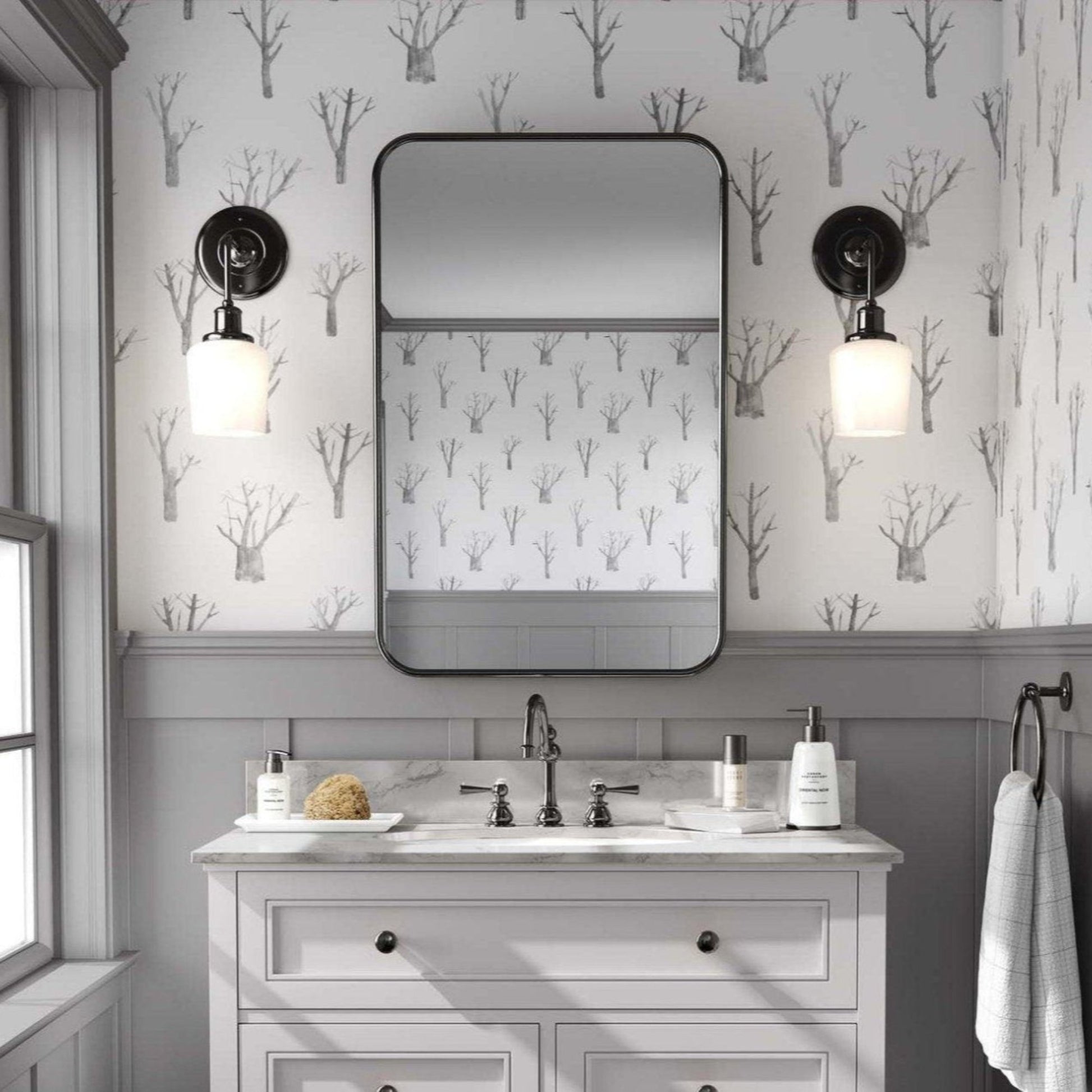 A classic powder room featuring grey wainscoting and minimalist wallpaper with a serene design of grey trees on a white background. The room is anchored by a sleek white vanity, complemented by a sophisticated grey and black mirror. Elegant sconces are placed symmetrically on either side, casting a warm, inviting glow.