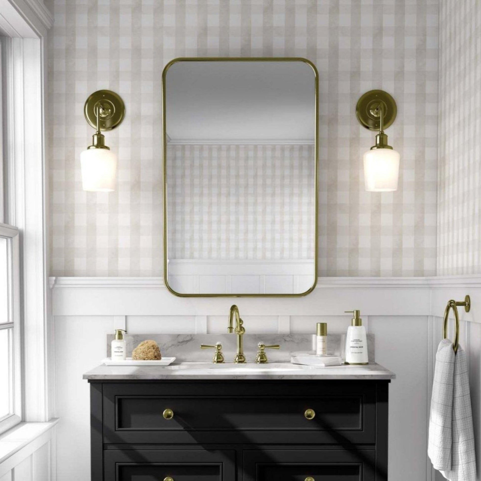 Bathroom with beige vichy wallpaper and a black vanity with gold accents. A large rectangular mirror with rounded edges hangs above the vanity, flanked by two brass sconces with white globe lights. 