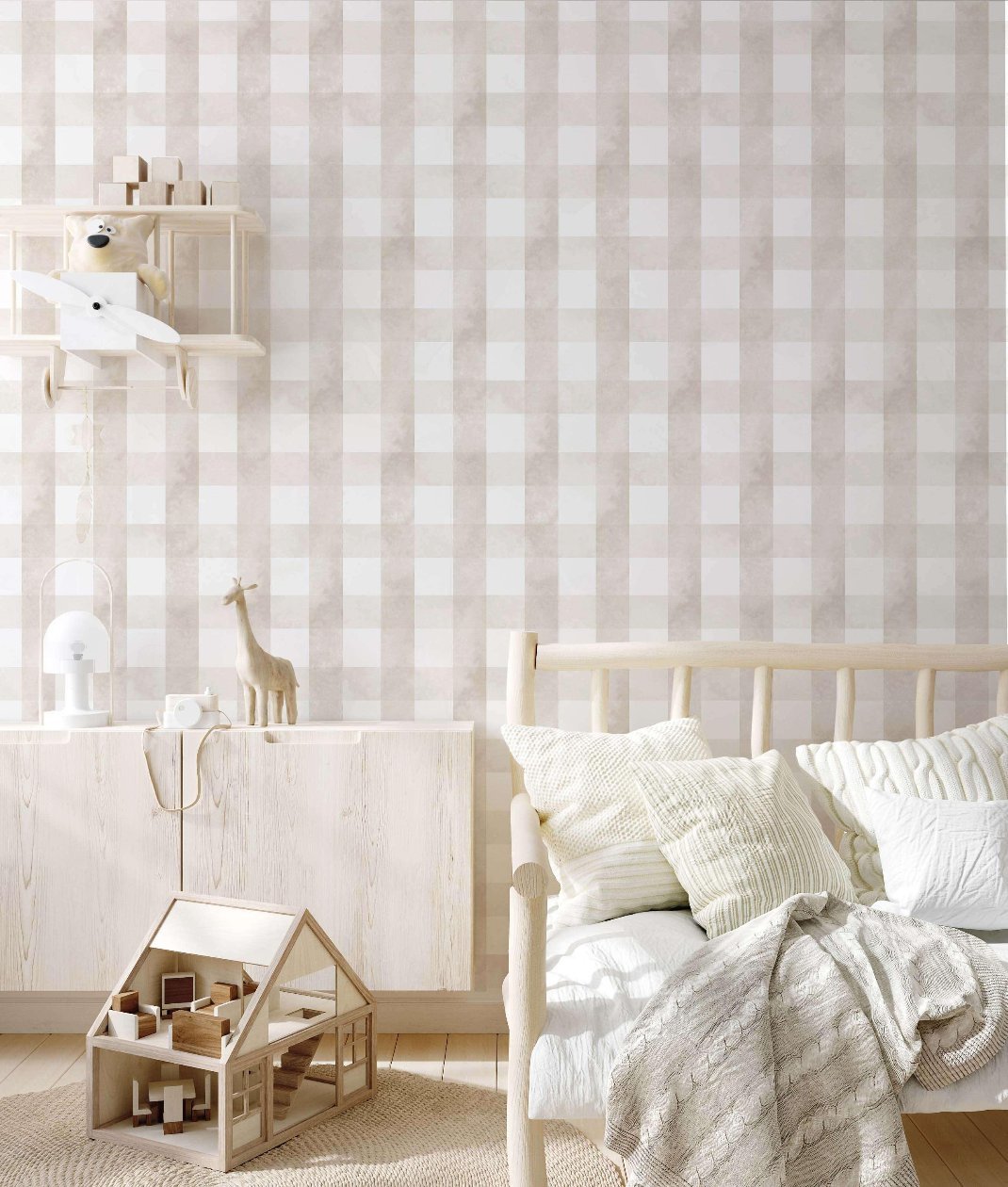 A serene children's bedroom with a light wooden bed, decorated with cream-colored pillows and a knitted blanket. A modern dollhouse is placed on the floor, next to a cabinet topped with a giraffe toy and a small lamp. A shelf above displays a toy airplane and a stuffed bear, set against beige vichy wallpaper.