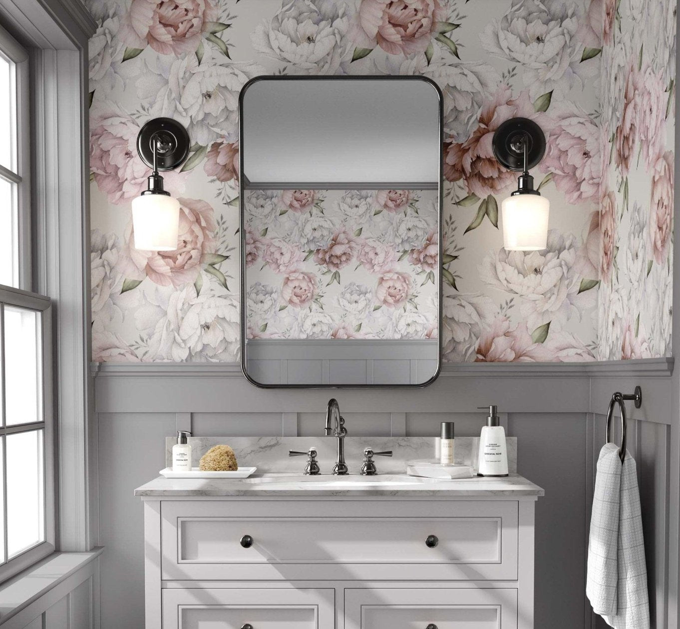 Bathroom with a light  pink and white peony floral wallpaper, featuring large flowers against a beige background. A white vanity with black fixtures stands below a rectangular mirror with rounded edges, flanked by brass sconces with globe lights. The space has an elegant, vintage feel, with grey paneling beneath the wallpaper and a window to the left allowing natural light.