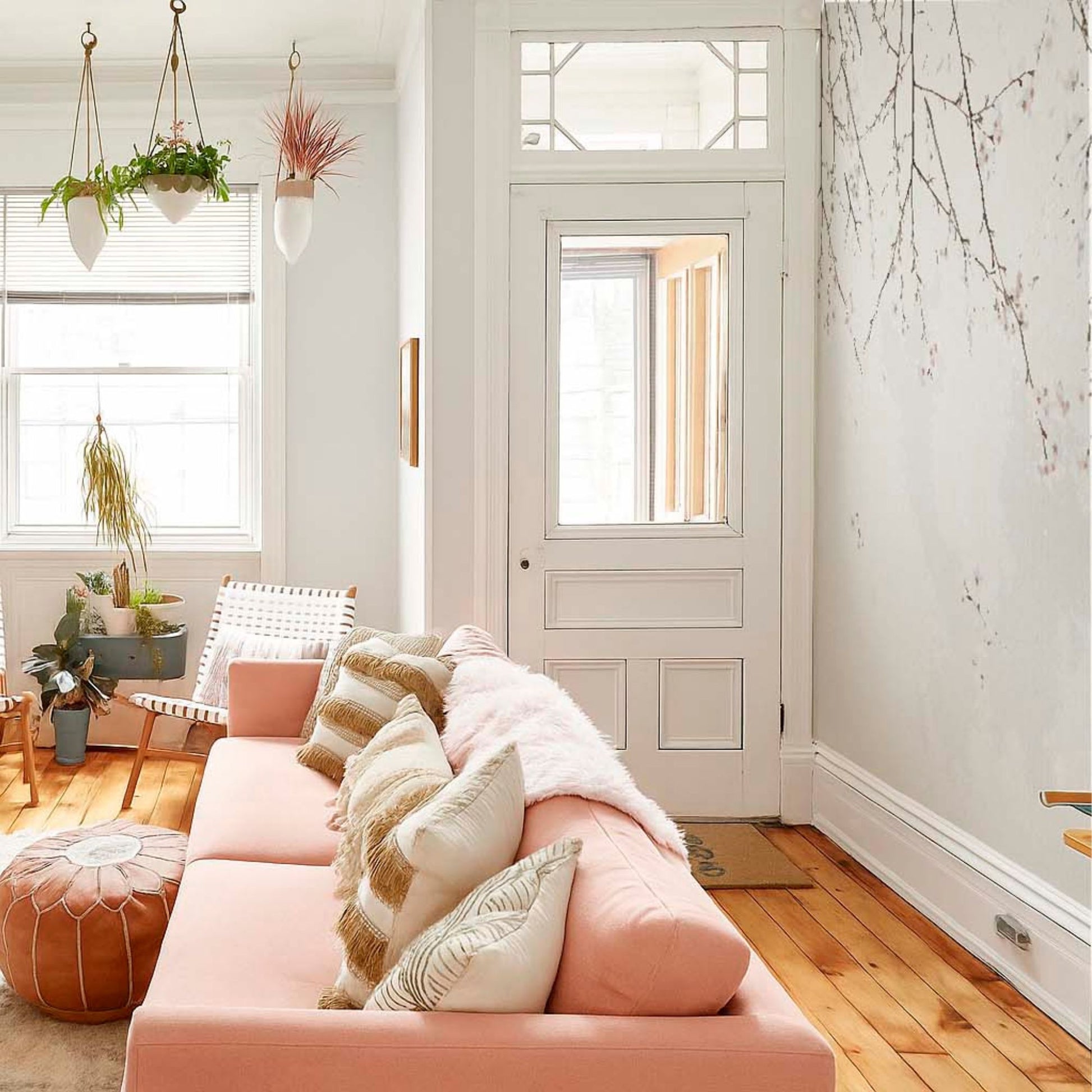 cat-610569683272 A bright, sunlit living room featuring a long pastel pink sofa adorned with striped and textured pillows, positioned against a large cherry blossom branch wallpaper mural and light hardwood floors.