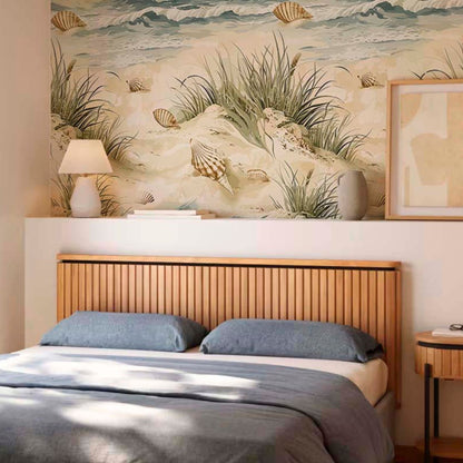cat-610567389512 A modern bedroom with a serene coastal aesthetic. A large wallpaper mural behind the bed depicts sand dunes, seagrass, and seashells in soft beige and green tones. Below the mural, a light wood slatted headboard sits behind a bed dressed in blue-grey pillows and a matching comforter. A white lamp, books, and a grey vase rest on a shelf above the bed.