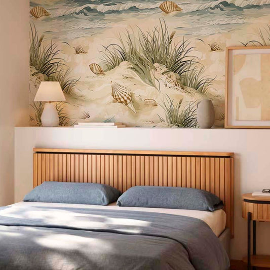 cat-610567389512 A modern bedroom with a serene coastal aesthetic. A large wallpaper mural behind the bed depicts sand dunes, seagrass, and seashells in soft beige and green tones. Below the mural, a light wood slatted headboard sits behind a bed dressed in blue-grey pillows and a matching comforter. A white lamp, books, and a grey vase rest on a shelf above the bed.