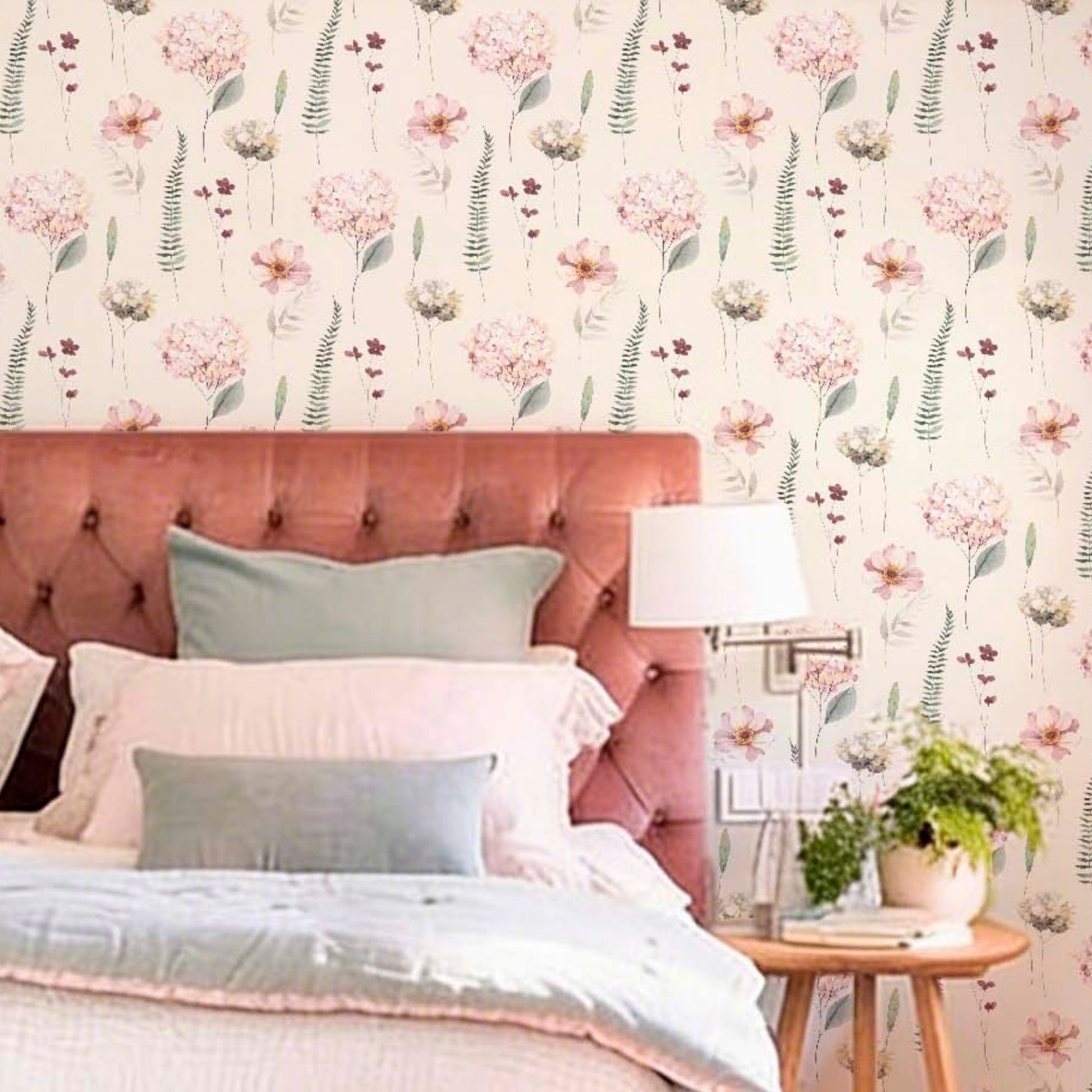 cat-616983200072 A bed with a pink tufted headboard and blue-grey pillows set against a floral hydrangea watercolor wallpaper. A wooden side table with a lamp and plant stands nearby.