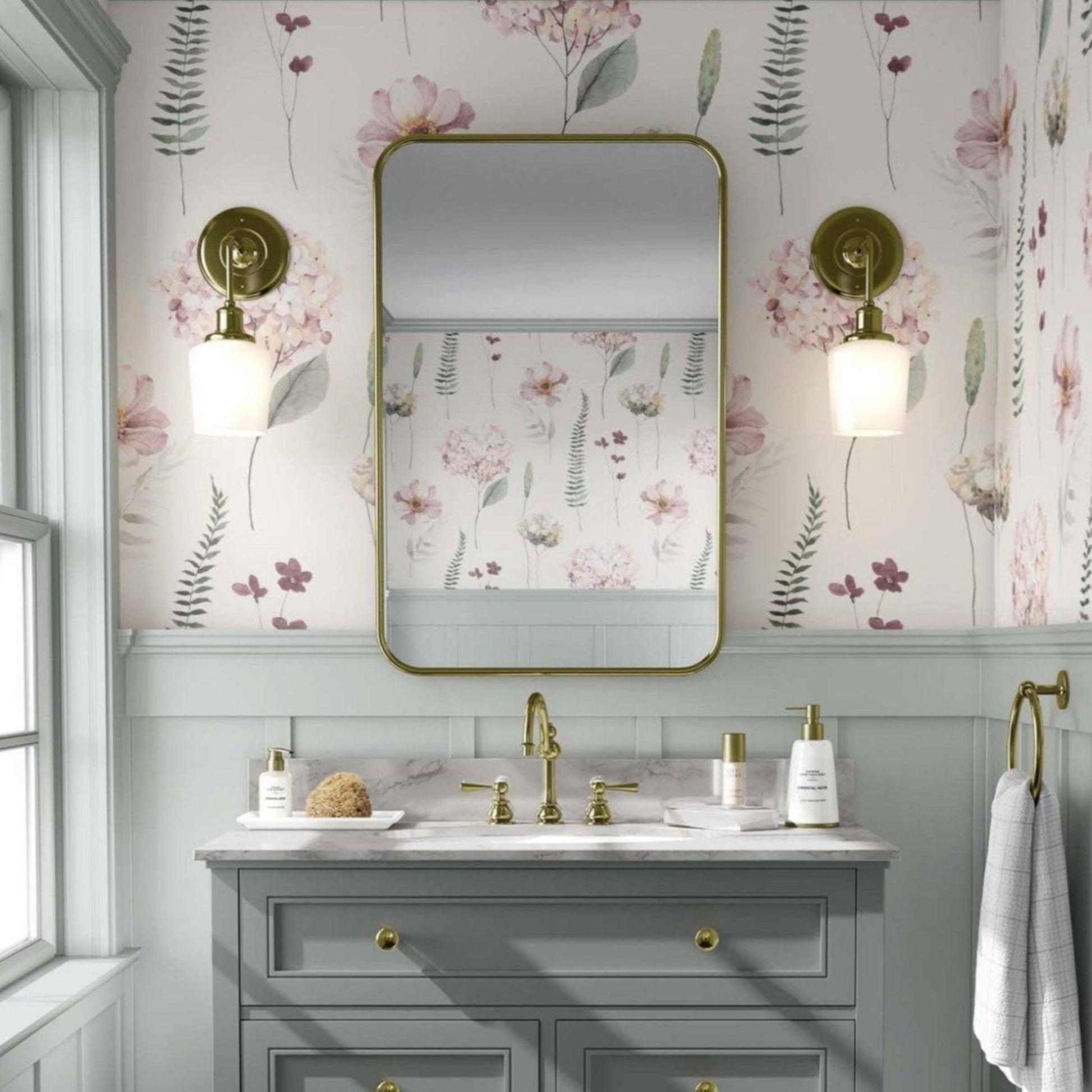 Bathroom with light  pink hydrangea flowers and greens floral wallpaper. A white vanity with gold fixtures stands below a rectangular mirror with rounded edges, flanked by brass sconces with globe lights. The space has an elegant, vintage feel, with light grey paneling beneath the wallpaper and a window to the left allowing natural light.