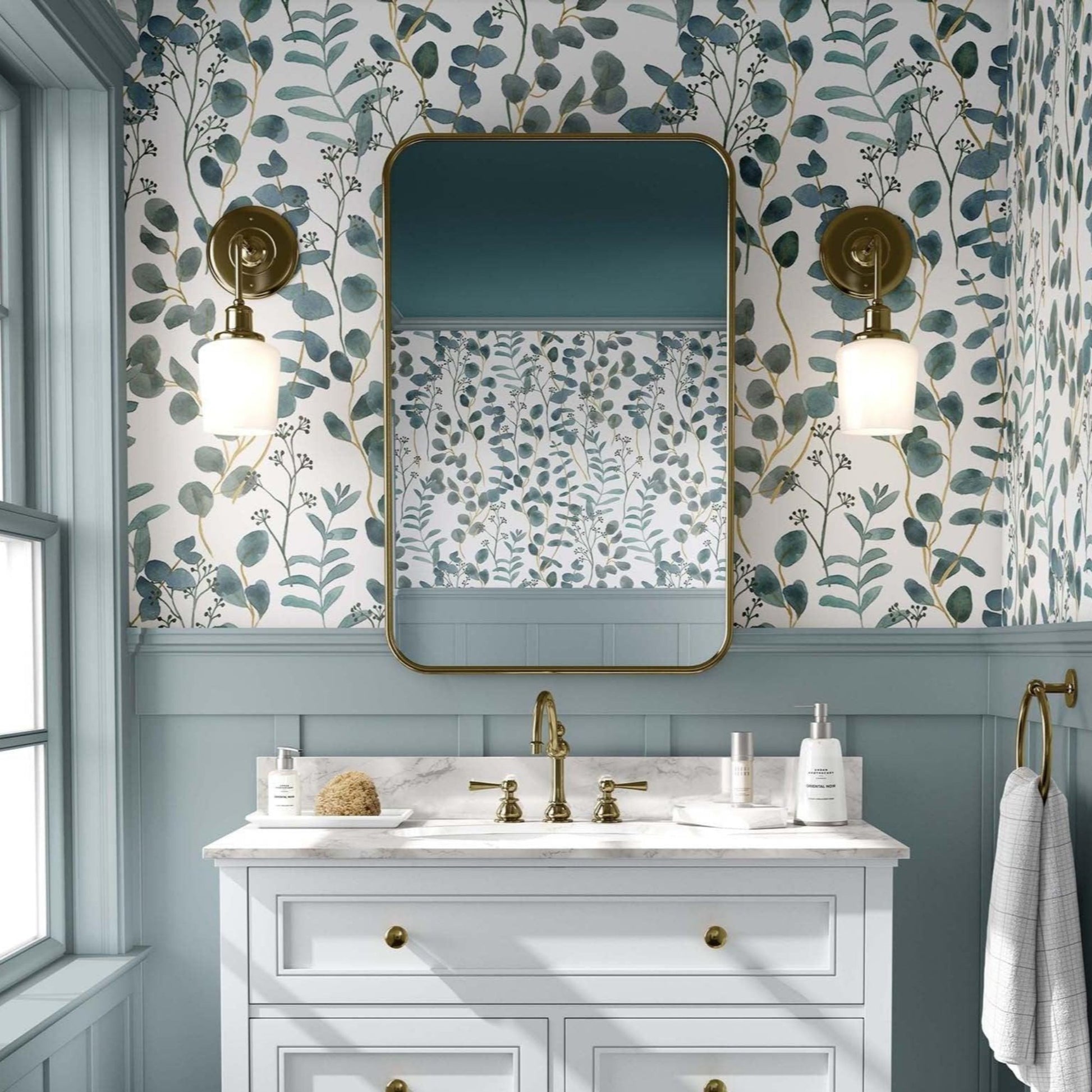 Bathroom with a green and white eucalyptus leaves against a white background. A white vanity with gold fixtures stands below a rectangular mirror with rounded edges, flanked by brass sconces with globe lights. The space has an elegant, vintage feel, with white paneling beneath the wallpaper and a window to the left allowing natural light.