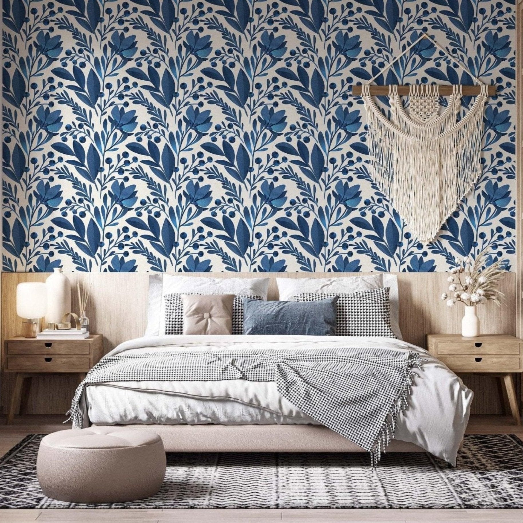 cat-616983200072 A modern bedroom featuring a bold blue and white botanical wallpaper as a central accent wall. The pattern consists of stylized navy and royal blue leaves and floral motifs. A large cream macramé wall hanging is centered above a bed with neutral-toned bedding, including a houndstooth throw and textured pillows. To the right, a light wood nightstand holds a white vase with dried flowers, complemented by light wood shelving and a minimalist aesthetic.
