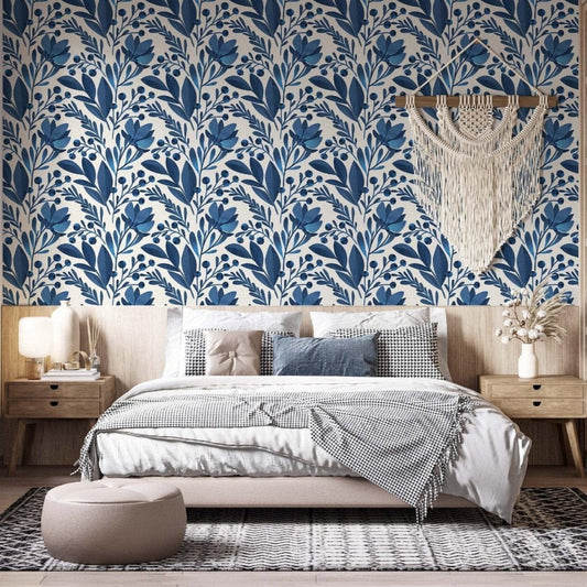 cat-616983200072 A modern bedroom featuring a bold blue and white botanical wallpaper as a central accent wall. The pattern consists of stylized navy and royal blue leaves and floral motifs. A large cream macramé wall hanging is centered above a bed with neutral-toned bedding, including a houndstooth throw and textured pillows. To the right, a light wood nightstand holds a white vase with dried flowers, complemented by light wood shelving and a minimalist aesthetic.
