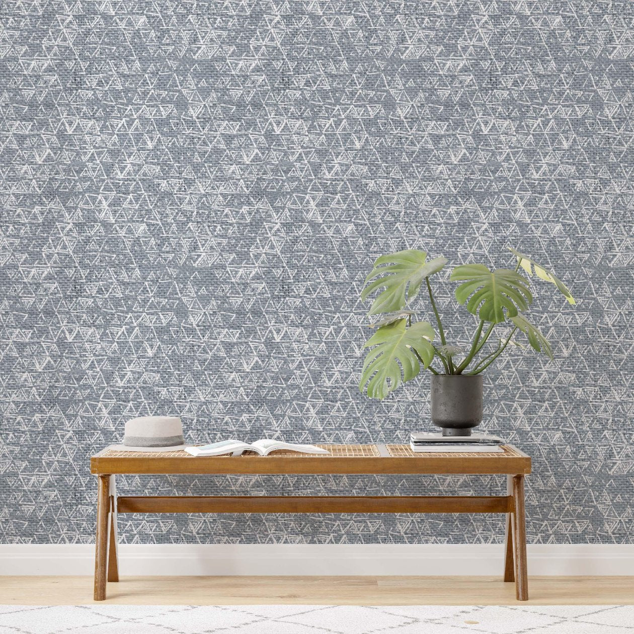 Close-up of a wall featuring a contemporary geometric wallpaper with a detailed, interlocking triangular pattern in a neutral grey color. The design adds texture and visual interest to the room, complemented by a wooden bench with an open book and a potted green monstera plant in a dark vase.