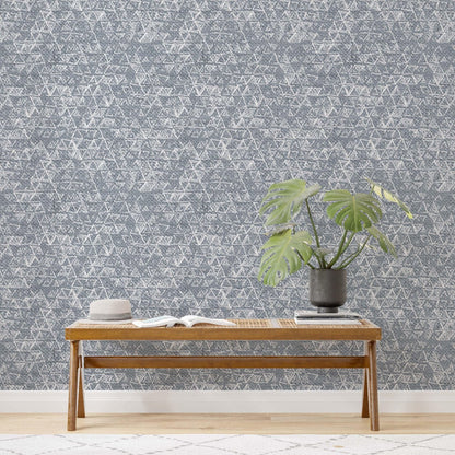 Close-up of a wall featuring a contemporary geometric wallpaper with a detailed, interlocking triangular pattern in a neutral grey color. The design adds texture and visual interest to the room, complemented by a wooden bench with an open book and a potted green monstera plant in a dark vase.