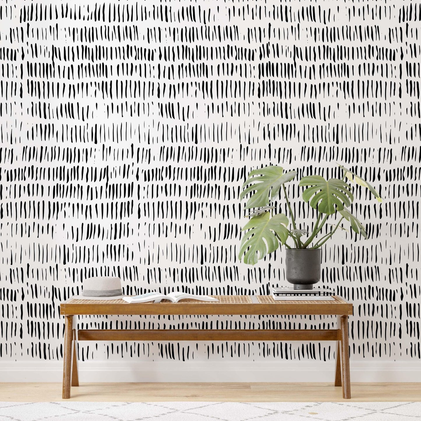 Close-up of a wall featuring a  black and white abstract wallpaper with irregular vertical strokes. The design adds texture and visual interest to the room, complemented by a wooden bench with an open book and a potted green monstera plant in a dark vase.