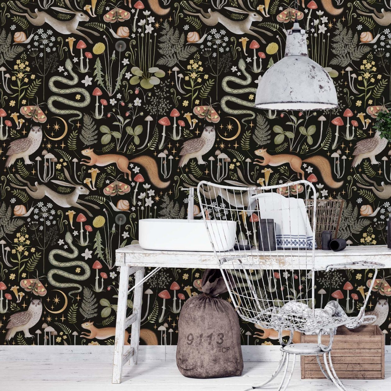 Room with rustic decor and a moody atmosphere, featuring a white table, wire chair, hanging metal lamp, and wallpaper with nature motifs—squirrels, snakes, and mushrooms—perfect for those who appreciate the cottagecore aesthetic.
