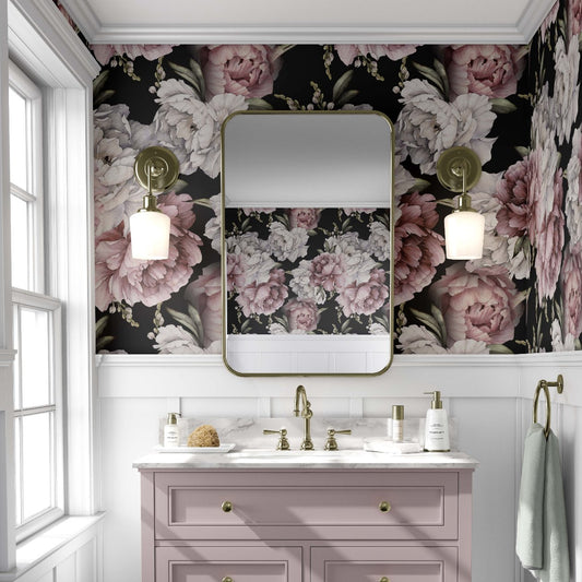 cat-610804236616 Bathroom with a bold pink and white peony floral wallpaper, featuring large flowers against a dark background. A light pink vanity with gold fixtures stands below a rectangular mirror with rounded edges, flanked by brass sconces with globe lights. The space has an elegant, vintage feel, with white paneling beneath the wallpaper and a window to the left allowing natural light.