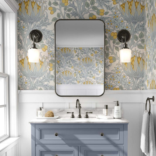 Bathroom with a pastel blue and yellow floral William Morris style wallpaper, featuring large Art Nouvau flowers against a beige background. A pastel blue vanity with black fixtures stands below a rectangular mirror with rounded edges, flanked by brass sconces with globe lights. The space has an elegant, vintage feel, with grey paneling beneath the wallpaper and a window to the left allowing natural light.