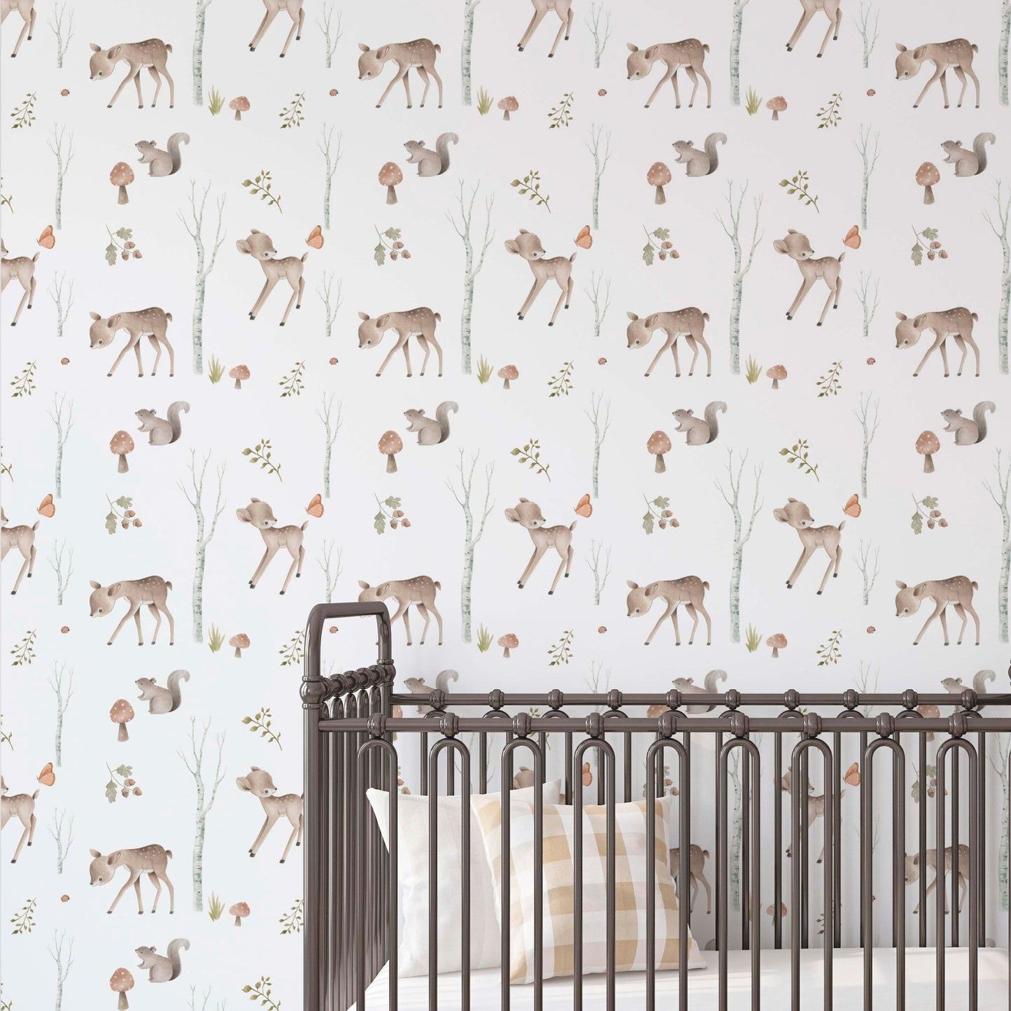 Nursery wall with woodland-themed wallpaper featuring illustrated fawns, squirrels, mushrooms, and birch trees, seen behind a dark metal spindle crib with a checkered pillow.