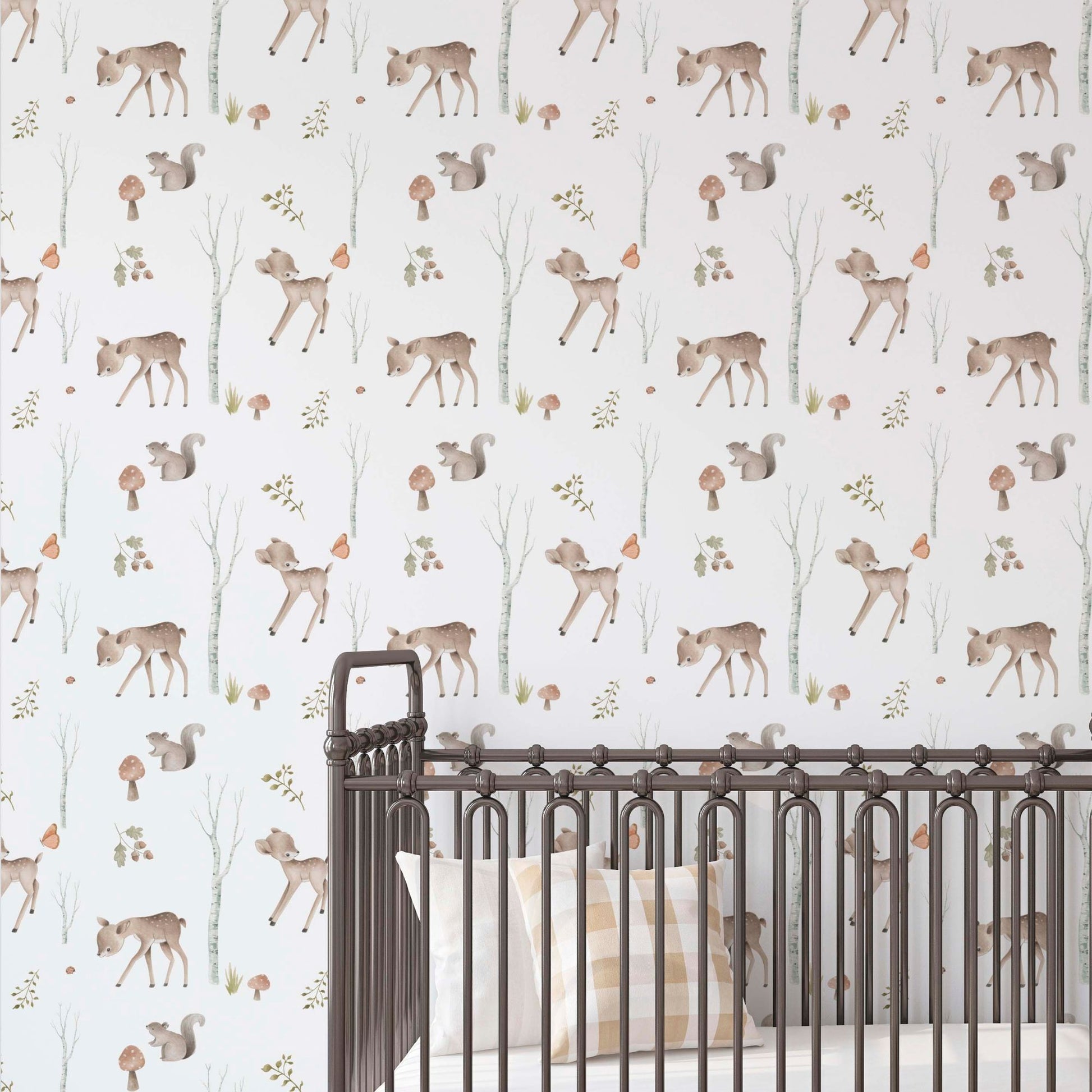 Nursery wall with woodland-themed wallpaper featuring illustrated fawns, squirrels, mushrooms, and birch trees, seen behind a dark metal spindle crib with a checkered pillow.