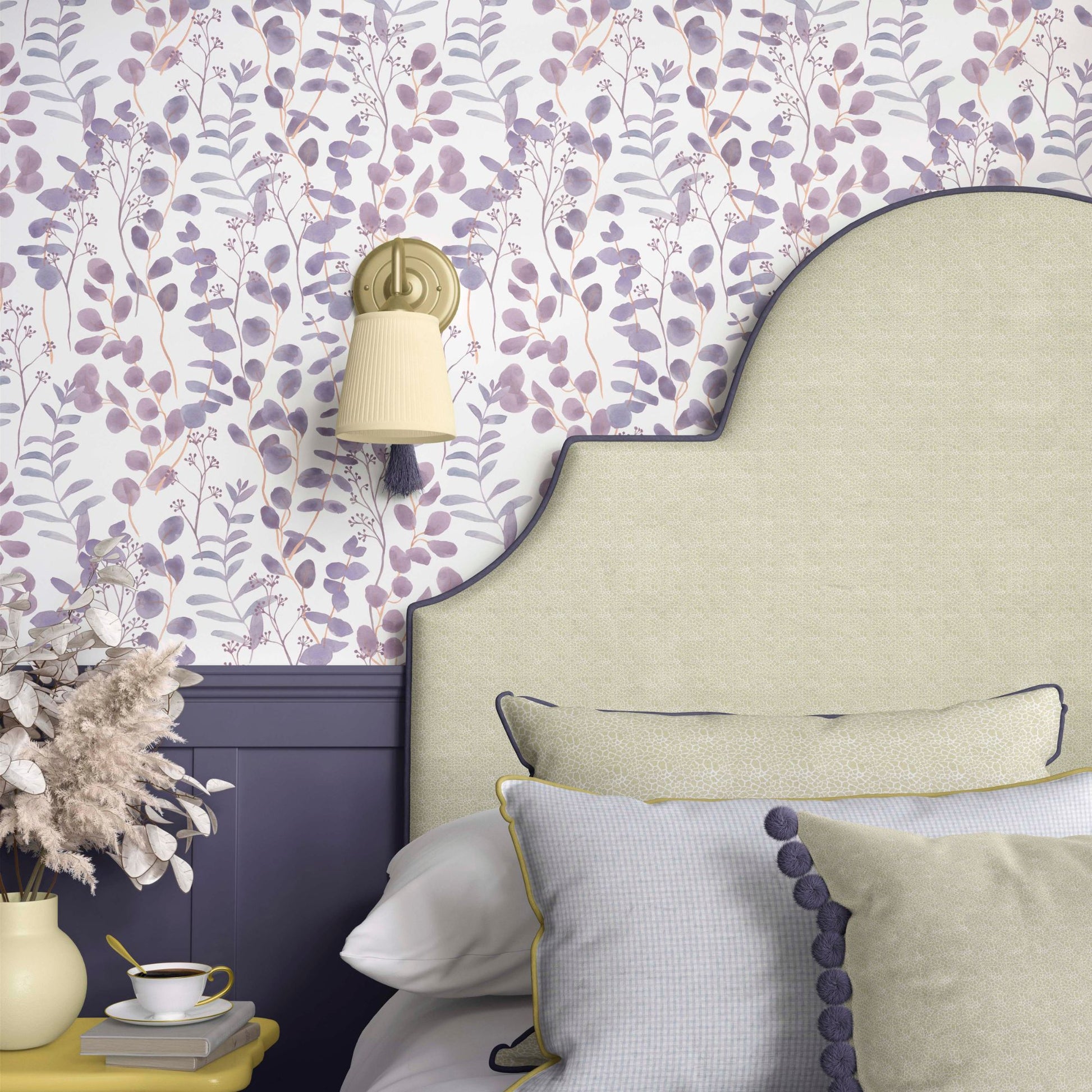 cat-616983200072 A close-up of a bedroom wall featuring lilac eucalyptus wallpaper above deep purple wainscoting. A beige upholstered headboard with purple piping is in front of the wall, with a small brass wall sconce with a pleated shade and tassel hanging above. A yellow bedside table with a teacup and a vase of dried pampas grass is partially visible