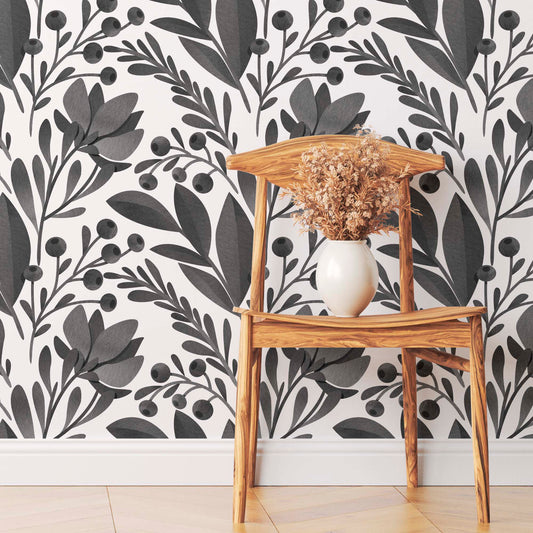 Bold black and white botanical pattern wallpaper featuring large leaves and berries behind a light wood chair and a vase with dried flowers.
