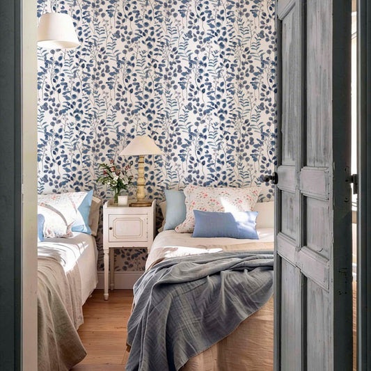 cat-615198294344 A cozy twin bedroom with a striking blue eucalyptus-style botanical wallpaper. The room features two single beds with neutral linen and blue accents, separated by a vintage white nightstand with a classic lamp. A rustic, grey-painted wooden door frames the view into the sunlit, serene space.