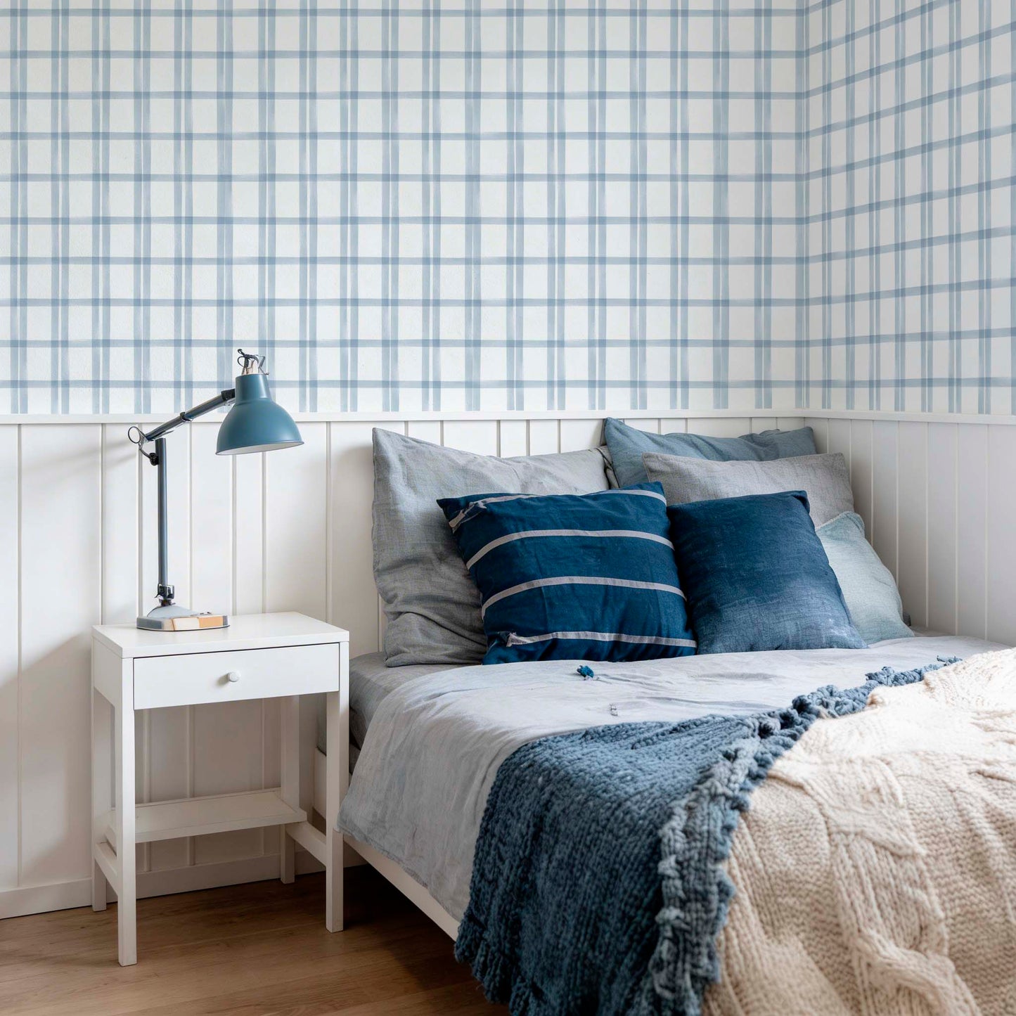 A cozy bedroom featuring a blue and white watercolor plaid wallpaper above white wood wall paneling. The bed is styled with blue and grey linens, a navy blue lamp on a white nightstand, and a knit throw blanket.