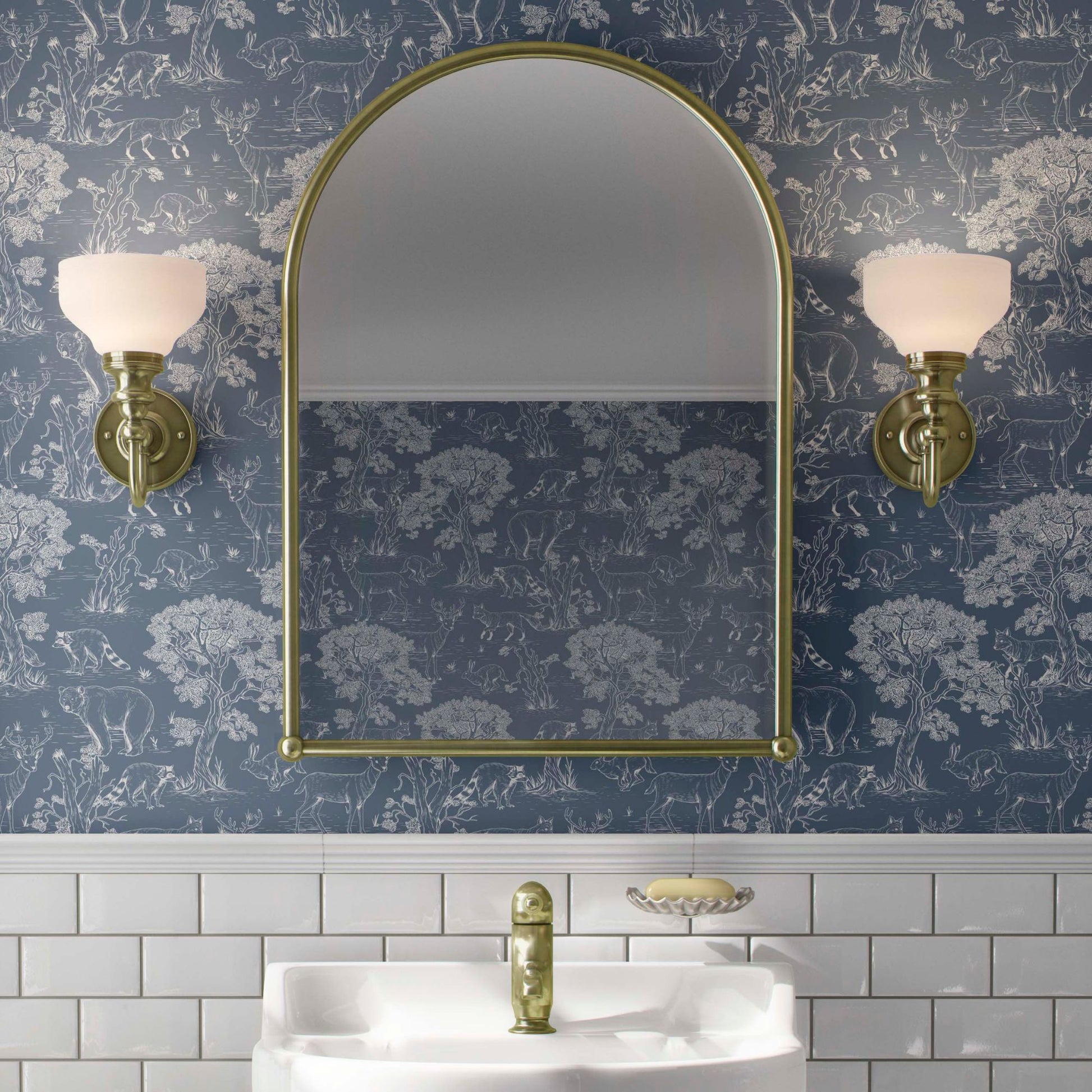 cat-610804236616 Elegant bathroom interior featuring blue woodland animals Toile wallpaper, a gold arched mirror, white subway tiles, and vintage brass wall sconces.
