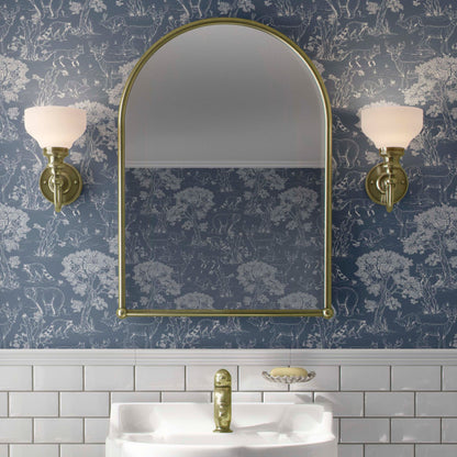 cat-610804236616 Elegant bathroom interior featuring blue woodland animals Toile wallpaper, a gold arched mirror, white subway tiles, and vintage brass wall sconces.
