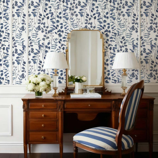 cat-610804433224 An elegant workspace featuring a blue and white botanical wallpaper with watercolor-style eucalyptus leaves and leafy branches. A classic wooden desk sits against the accent wall, topped with a gold-framed mirror and two traditional table lamps with white pleated shades. In the foreground, a matching wooden chair with blue and white striped upholstery completes the sophisticated, traditional aesthetic.
