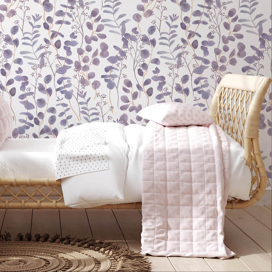 cat-615198294344 A bright children's bedroom featuring a white wall covered in lilac eucalyptus leaf wallpaper. In front of the wall, there is a light-colored rattan daybed with pink and white bedding, a large plush teddy bear sitting on a round jute rug, and a matching rattan chair to the left.