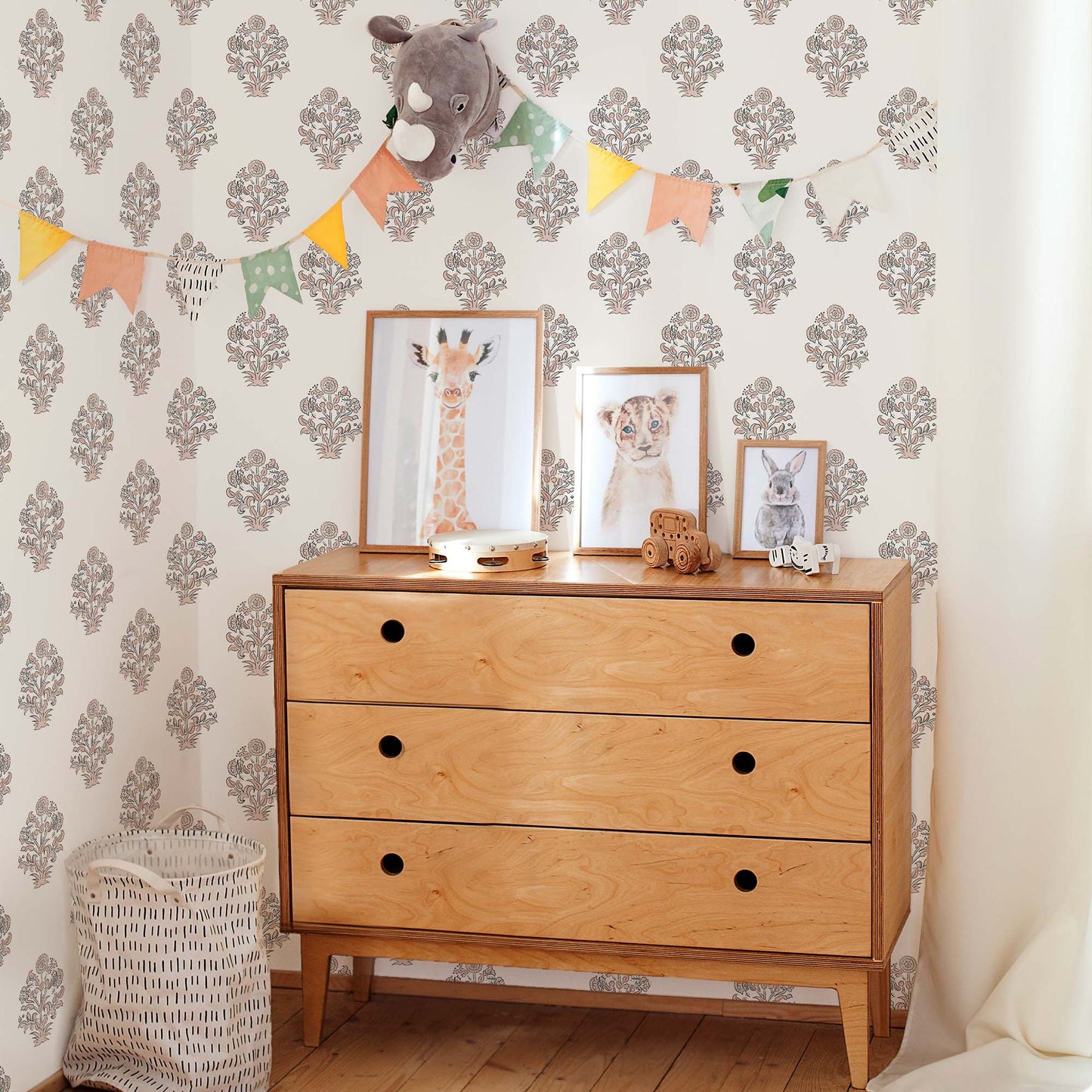 A serene and organized gender-neutral nursery featuring a charcoal floral block print wallpaper with traditional buta motifs. A mid-century modern wooden dresser with circular cut-out handles sits against the accent wall, topped with framed watercolor animal prints of a giraffe, lion, and rabbit. A colorful triangle bunting garland and a plush rhinoceros wall mount add a whimsical and playful touch to the sophisticated space.
