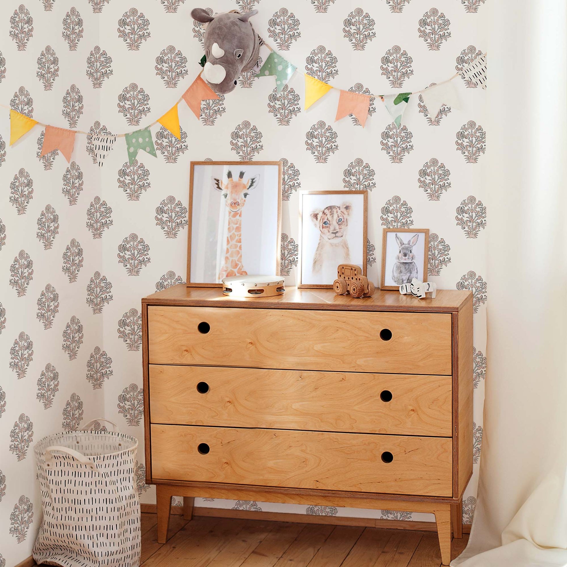 A serene and organized gender-neutral nursery featuring a charcoal floral block print wallpaper with traditional buta motifs. A mid-century modern wooden dresser with circular cut-out handles sits against the accent wall, topped with framed watercolor animal prints of a giraffe, lion, and rabbit. A colorful triangle bunting garland and a plush rhinoceros wall mount add a whimsical and playful touch to the sophisticated space.