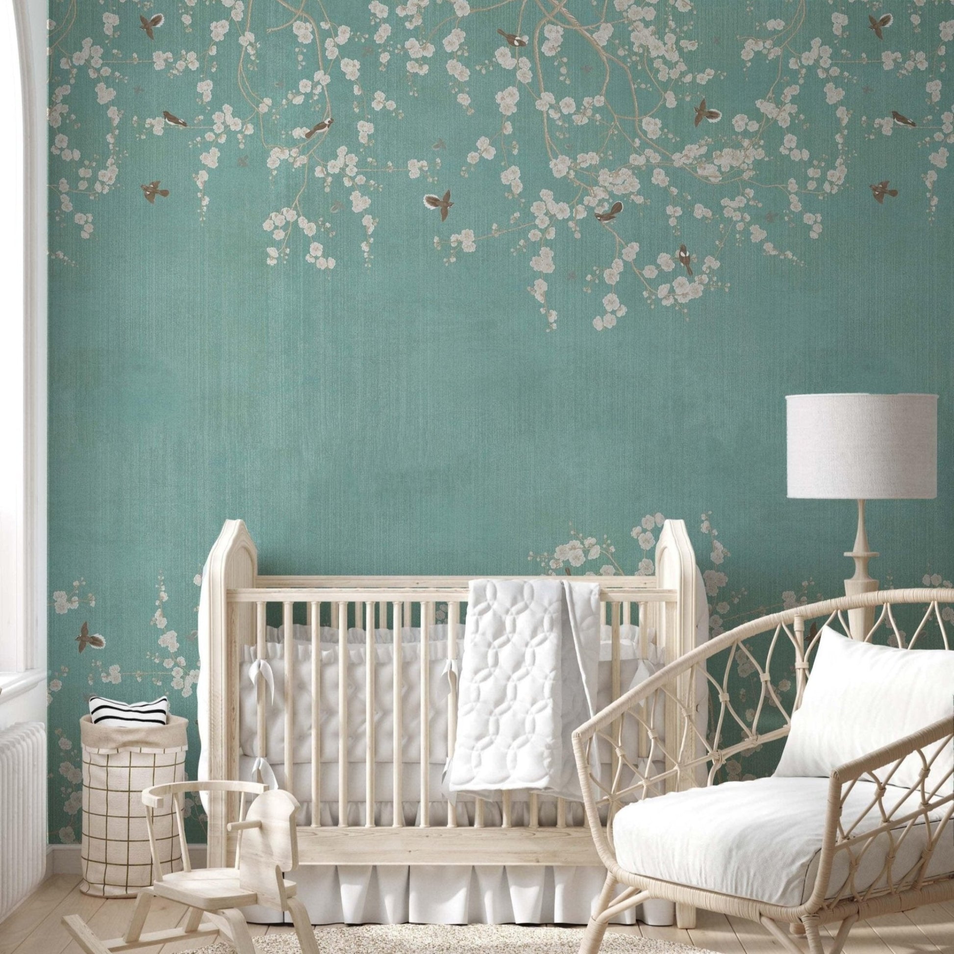 cat-610568044872 Nursery room featuring a teal Chinoiserie wallpaper mural with white cherry blossoms and small birds, styled with a white wooden crib and a wicker rocking chair.