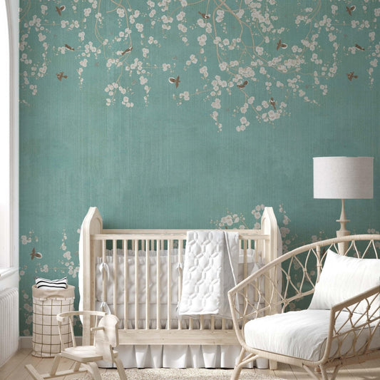 cat-610568044872 Nursery room featuring a teal Chinoiserie wallpaper mural with white cherry blossoms and small birds, styled with a white wooden crib and a wicker rocking chair.